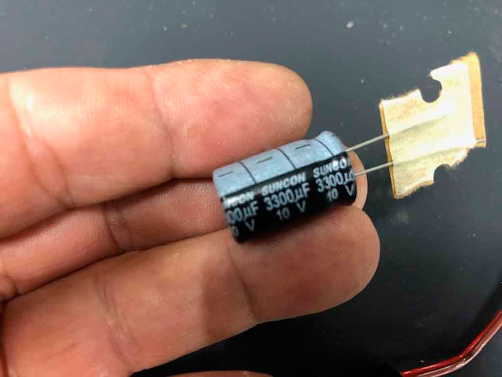 Close up of a perfectly run-of-the-mill 3300?f 10V capacitor, with the white stripe marking the negative (-) leg held in a hand.