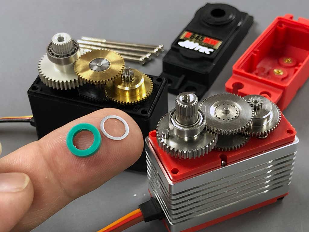 Extreme close up of the Viton seal and packing washer for the splined output shaft of the ProModeler DS180DLHV placed on a fingertip in the foreground against the exposed gear trains of both a Hitec D645MW and ProModeler DS180DLLV standard class servos against a neutral gray background.