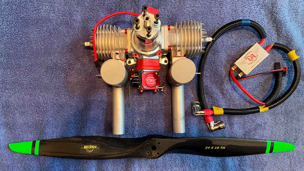 Close up of Desert Aircraft DA70 twin cylinder engine with ignition module and propeller arrayed beside it on a light blue towel.