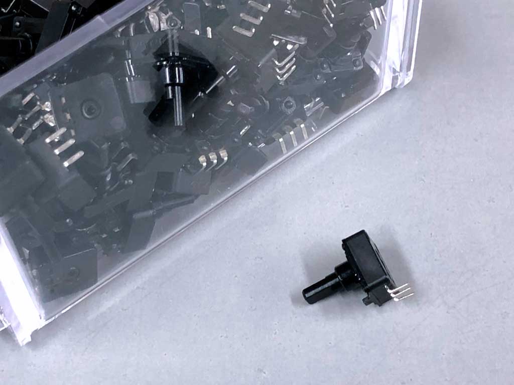 Close up of clear plastic bin with Nobel 1mc million cycle potentiometers and a single capacitor laying on the table before it.