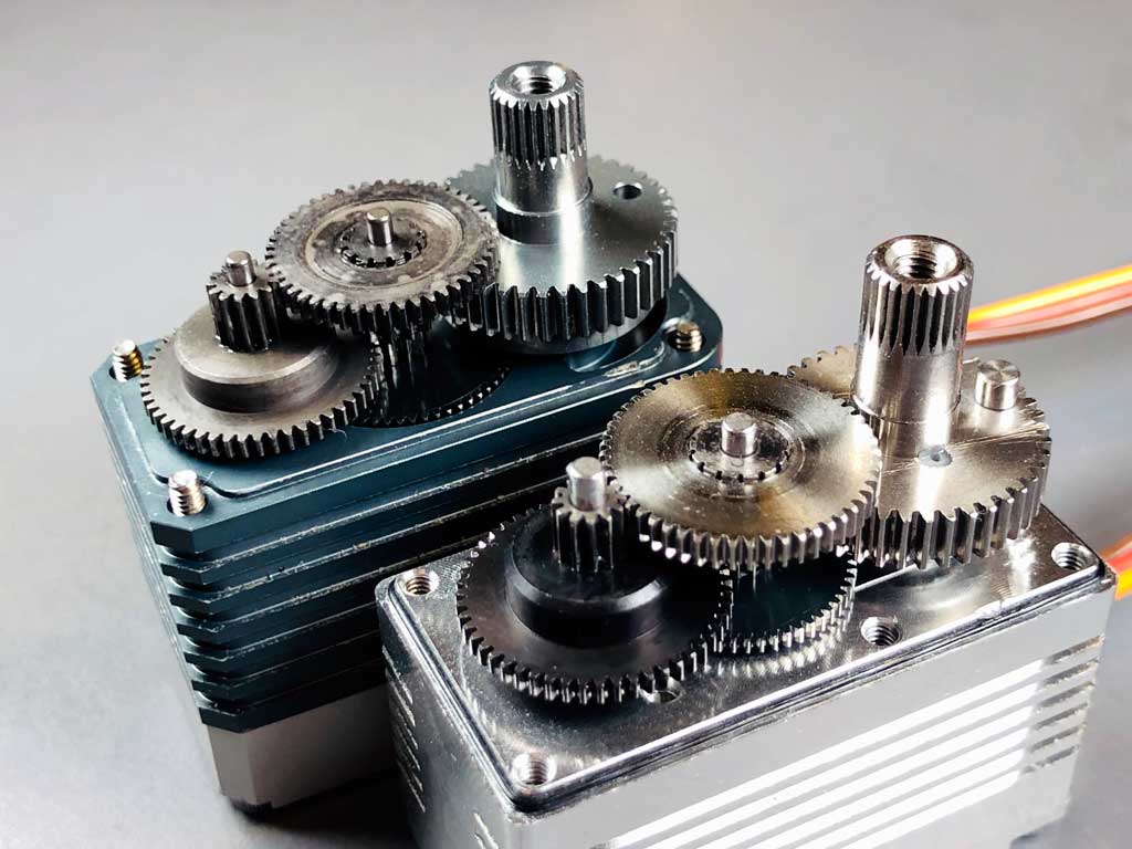 Close up of the gear train of a pair of standard class brushless servos with equipped with steel gears. The principal difference being the ProModeler servo uses a more durable stainless steel output gear. This is easy to see, also as the soft sheen of 303 stainless bull gears and 412 stainless pinion output gears give the ProModeler the edge.